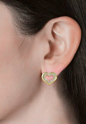 Orecchini a forma di cuore in oro con un centro vuoto, contornati da piccoli cristalli trasparenti, indossati all'orecchio di un modello.
