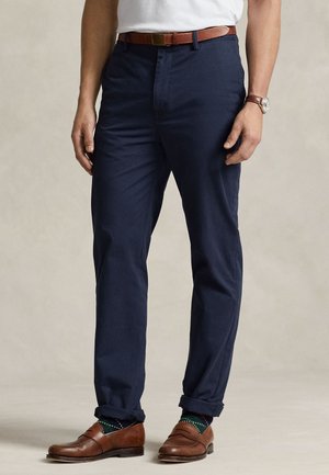 Homme portant un pantalon bleu marine sur mesure avec une ceinture en cuir marron et des mocassins marron assortis, associés à des chaussettes vertes à motifs et une chemise blanche.