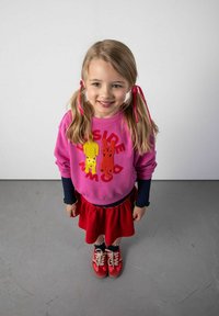 Jasno różowa bluza z kolorowymi aplikacjami zwierząt, czerwona spódniczka z falbanami, granatowa bluzka z długim rękawem oraz czerwone trampki.