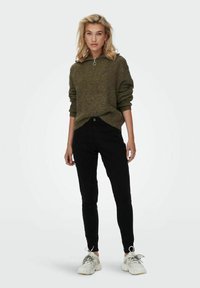 Pull en maille vert olive avec un col haut et une fermeture éclair argentée, associé à un jean skinny noir et des baskets blanches avec des accents gris.