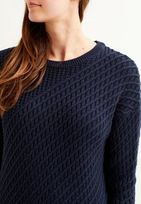 Maglione blu navy lavorato a maglia con un motivo a diamante testurizzato, scollo a girocollo e maniche lunghe. Il tessuto sembra morbido e caldo.