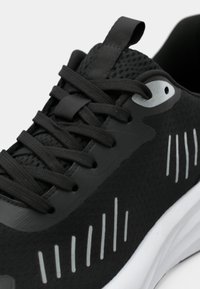 Fekete sportcipő texturált hálós felsőrésszel, lapos fűzőkkel és fényvisszaverő részletekkel. Kerek fűzőlyuk kialakítással és fehér talppal.