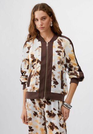 Woman with long wavy hair wearing a white and brown floral zip-up jacket and matching pants, posing with silver bracelets on left wrist.