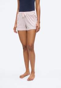 Γυναίκα που φορά ριγέ σορτς με κορδόνι σε ανοιχτό ροζ και ναυτικό μπλε χρώμα και αμάνικη μπλούζα σε ναυτικό μπλε, στεκόμενη γυμνόποδη σε μονόχρωμο φόντο.