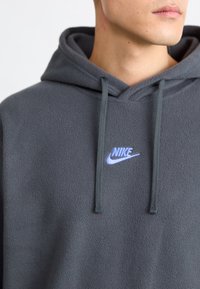 Grå fleece-hoodie med justerbar huva och en liten blå Nike-logotyp på bröstet. Mjuk textur och avslappnad passform.
