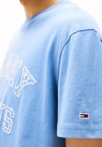 Camiseta de algodón de color azul claro con texto gráfico blanco en el frente y un parche de logo en rojo, blanco y azul en la manga. Manga corta.