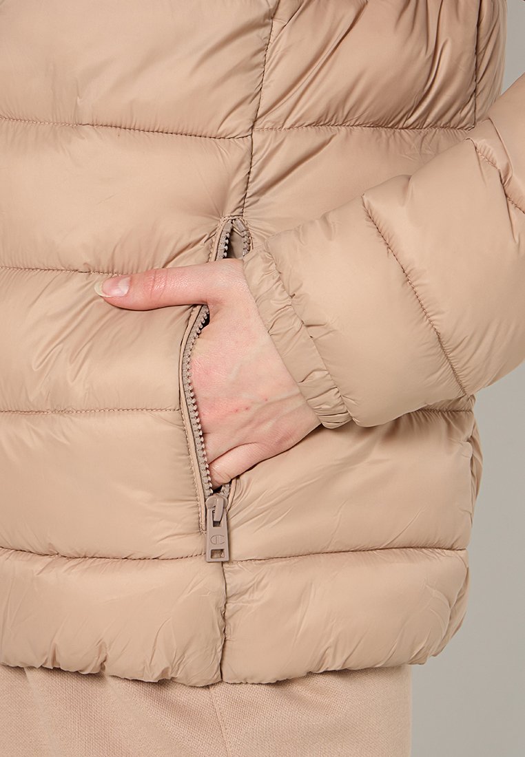 Giacca imbottita beige con tasca con zip, texture gonfiata e polsini elasticizzati. Il tessuto ha una finitura liscia con dettagli di cucitura visibili.