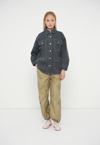 Veste en denim foncé avec fermeture à boutons et deux poches poitrine, associée à un pantalon cargo beige clair. Comprend des poignets zippés et élastiques. Chaussures roses.