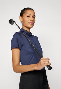 Polo shirt blu navy con maniche corte, caratterizzato da una patta con bottoni. Una golfista tiene una mazza nera sulla spalla, con impugnature texturizzate visibili.