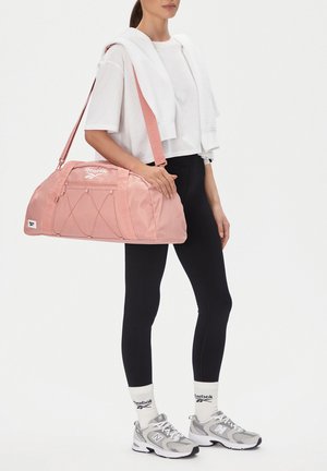 Osoba ubrana w białą krótka koszulkę, czarne legginsy, białe skarpetki Reebok, szare trampki, niosąca różową torbę sportową Reebok na ramieniu.