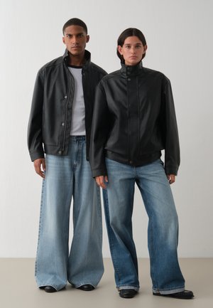 Deux modèles posent dans un studio, vêtus de vestes en cuir noires et de jeans bleus amples.