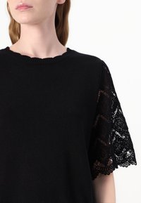 Černý pletený top se scalloped výstřihem a krajkovými krátkými rukávy, zdobený složitými květinovými vzory, hladkým materiálem a volným střihem.
