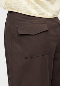 Κοντινή εικόνα σκούρου καφέ παντελονιού με μεγάλη τσέπη με καπάκι στο πλάι, συνδυασμένο με υφασμάτινο εκρού τοπ.