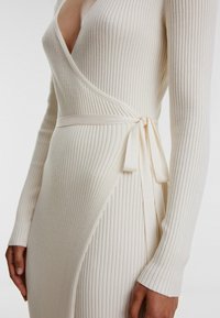 Rippiertes cremefarbenes Wickelkleid mit langen Ärmeln und einem Bindeband in der Taille. Verfügt über einen tiefen Ausschnitt und einen strukturierten Stoff.