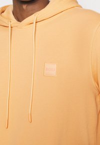 Sweat à capuche orange clair avec cordon de serrage, arborant une petite étiquette carrée avec le logo "BOSS". Texture de tissu lisse et détails de couture contrastants.