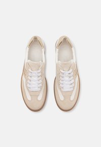 Béžové semišové a bílé kožené tenisky s kulatou špičkou, polstrovaným límcem, bílými tkaničkami a hnědými akcenty po stranách a na podrážce.