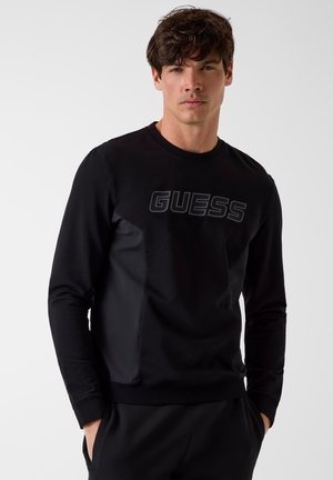 Jeune homme portant un sweat-shirt noir à manches longues avec le logo « GUESS », associé à un pantalon noir, posant les mains dans les poches sur un fond blanc.