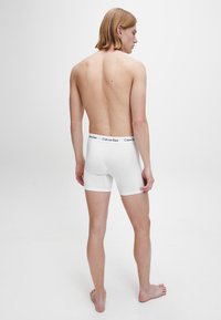 Bijele pamučne bokserice s crnim pojasom Calvin Klein, prilagođenog dizajna, prikazuju stražnji pogled osobe.