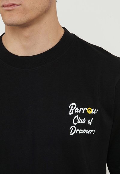 Man wearing black crew neck shirt with white text "Barrow Club of Dreamers" and yellow smiley face on the left chest.