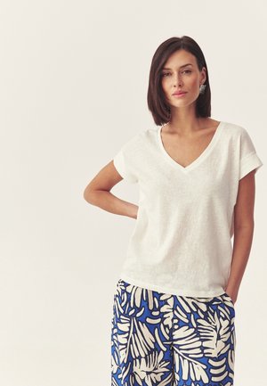 Woman with short brown hair wearing white V-neck short sleeve top and blue and beige patterned pants, standing with one hand on hip.