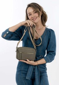 Sac croisé gris texturé avec une poche zippée à l'avant et un logo, doté une longue bandoulière ajustable de couleur assortie.