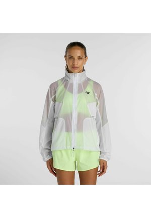 Frau trägt eine weiße, durchsichtige Reißverschlussjacke über einem neon-gelben Sport-BH und passenden Shorts, steht vor einem schlichten Hintergrund.