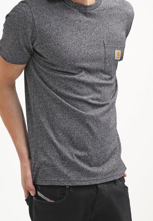 Hombre que lleva una camiseta gris texturizada de manga corta con un bolsillo delantero y pantalones negros, de pie con una mano en el bolsillo.