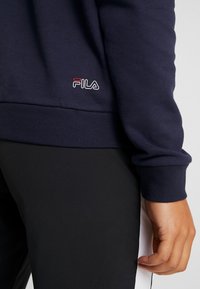 Felpa blu navy con polsini e orlo a coste, caratterizzata da un piccolo logo Fila rosso e bianco sul davanti, abbinata a pantaloni neri.