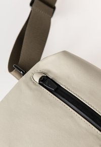Borsa beige con una texture liscia, dotata di una cerniera nera e una tracolla in tessuto marrone. Design compatto adatto per l'uso quotidiano.