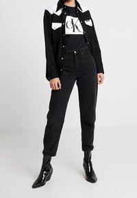 Schwarze Jeans aus Denim, kombiniert mit einem schwarzen Shirt mit weißen Akzenten und einem Logo-T-Shirt. Glänzende schwarze Stiefeletten. Lässiges Outfit-Design.