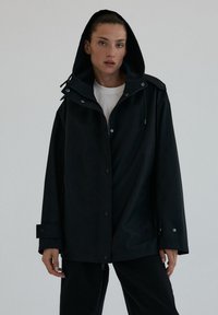 Kobieta w czarnej, oversize'owej kurtce z kapturem, nosząca białą koszulę, stoi na jednolitym jasnym tle z neutralnym wyrazem twarzy.