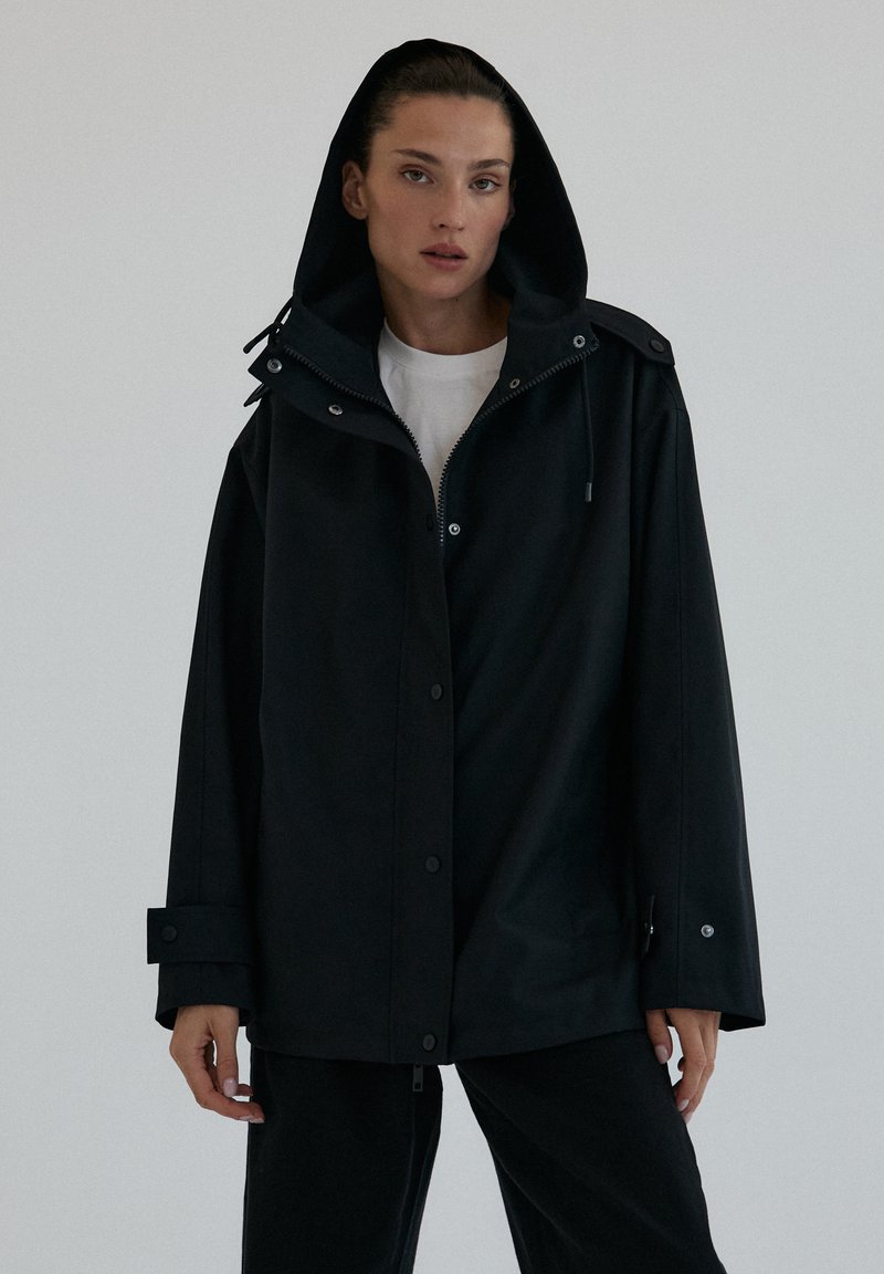 Kobieta w czarnej, oversize'owej kurtce z kapturem, nosząca białą koszulę, stoi na jednolitym jasnym tle z neutralnym wyrazem twarzy.