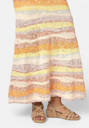 Piernas inferiores con sandalias de cuña de ante beige con tiras, bajo una falda larga de punto con rayas horizontales en tonos pastel naranja, amarillo y beige.