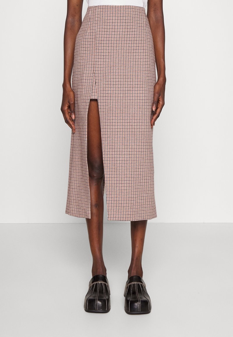 Femme portant une jupe midi à carreaux beige et rose avec une fente devant, associée à des chaussures noires à bout carré et coutures visibles.