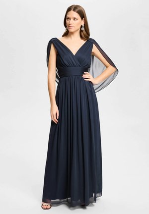 Femme portant une robe bleu marine longue jusqu'au sol avec une jupe plissée, un décolleté en V, des manches cape transparentes, posant avec une main sur la hanche.