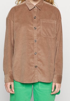 Widok z przodu osoby noszącej brązową sztruksową koszulę na guziki z kieszenią na piersi oraz jaskrawozielone spodnie na neutralnym tle.