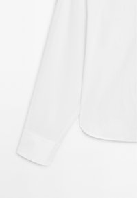 Langärmliges weißes Hemd aus Baumwolle. Verfügt über einen geraden Kragen und Knopfmanschetten. Glatte Textur mit einem leicht abgerundeten Saum.