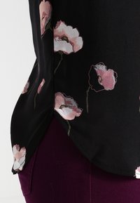 Černá květinová blůza s růžovými mákovými vzory, hladký materiál, mírně zakřivený lem, spárovaná s tmavě fialovými kalhotami.