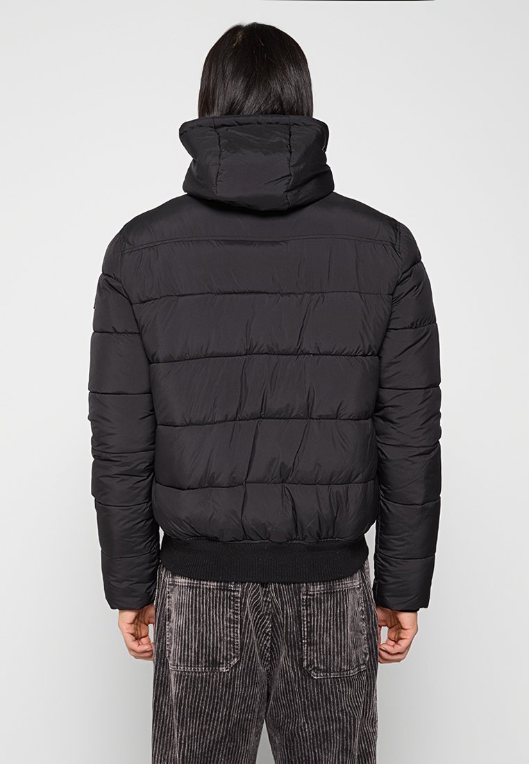 Man in een zwart gewatteerd jack met hoge kraag en grijze corduroy broek, gezien van achteren tegen een effen achtergrond.