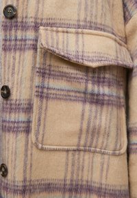Gros plan d’un manteau en laine beige avec un motif à carreaux violets, une poche à rabat et des boutons foncés sur le côté gauche.