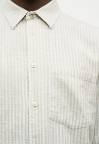 Chemise beige clair à boutons avec fines rayures verticales, dotée d'un col pointu et d'une poche poitrine unique. Tissu texturé.