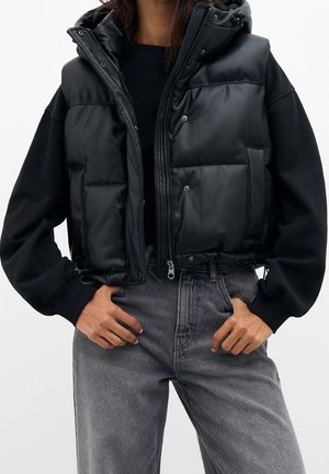 Osoba ubrana w czarną pikowaną kamizelkę na napy i zamek błyskawiczny, na czarnej bluzie oraz szare, dopasowane jeansy z wysokim stanem, z rękami w przednich kieszeniach.