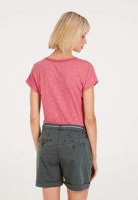 Blusa rosa a maniche corte con maniche arrotolate, abbinata a pantaloni corti verde scuro con orlo arrotolato, dettagli con bottoni e tasche posteriori.