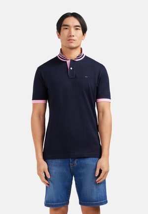 Homme portant un polo bleu marine avec col et poignets roses, associé à un short en denim bleu, sur fond blanc.