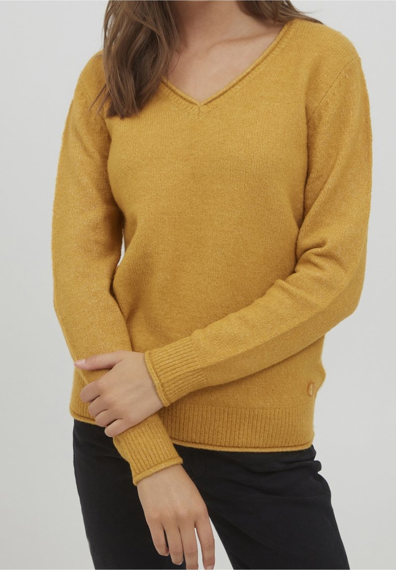 Femme portant un pull en maille jaune moutarde à col en V et un pantalon noir, les bras croisés à la taille sur un fond uni.