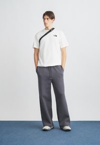 Λευκό βαμβακερό T-shirt με μαύρο λογότυπο, συνδυασμένο με γκρι, φαρδιά φόρμα. Τα παπούτσια είναι ανοιχτό γκρι με σκούρες λεπτομέρειες. Ρυθμιζόμενη λεπτομέρεια στο λουράκι.