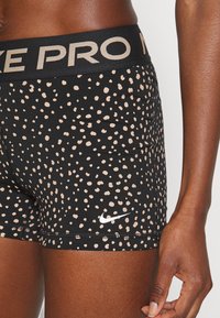 Pantalones cortos deportivos negros con un patrón moteado en beige, que cuentan con una amplia cinturilla negra con "NIKE PRO" en dorado y un logo de Nike en blanco.