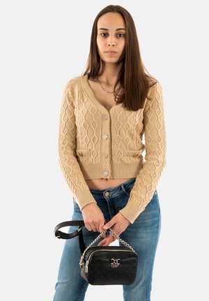 Cardigan beige a maglia con motivo a diamante testurizzato, abbinato a jeans blu scuro. Tiene in mano una piccola borsa nera con manico a catena e logo.
