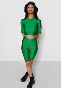 Conjunto de activewear verde de duas peças com mangas curtas e calções de cintura alta, com detalhes em vermelho e um corte. Botas pretas usadas.