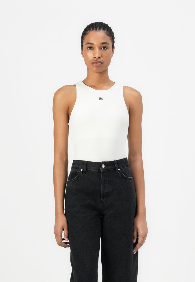 Hvit ermeløs bodysuit med rund hals og frontlogo, kombinert med høyhåndet svarte jeans, som fremhever en tettsittende silhuett.